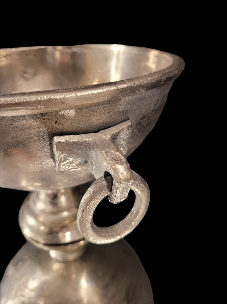 May include: Close-up of a silver-colored metal chalice with a rounded bowl and a ring handle. The chalice sits on a pedestal base, reflecting the light. The background is black.