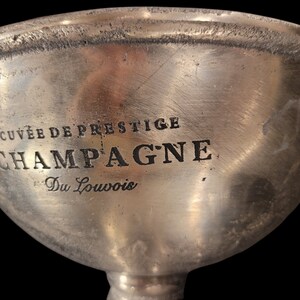 May include: A silver champagne bucket with the words "CUVEE DE PRESTIGE CHAMPAGNE Du Louvois" etched into the side. The bucket has two small handles and a wide, shallow bowl. The background is black.