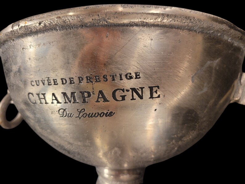 May include: A silver champagne bucket with the words "CUVEE DE PRESTIGE CHAMPAGNE Du Louvois" etched into the side. The bucket has two small handles and a wide, shallow bowl. The background is black.