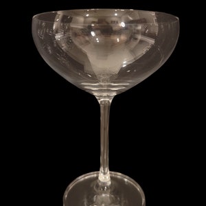 May include: A clear, stemmed champagne coupe glass with a wide, shallow bowl and a thin stem. The glass sits on a round base. The glass reflects the surrounding environment, with a black background.