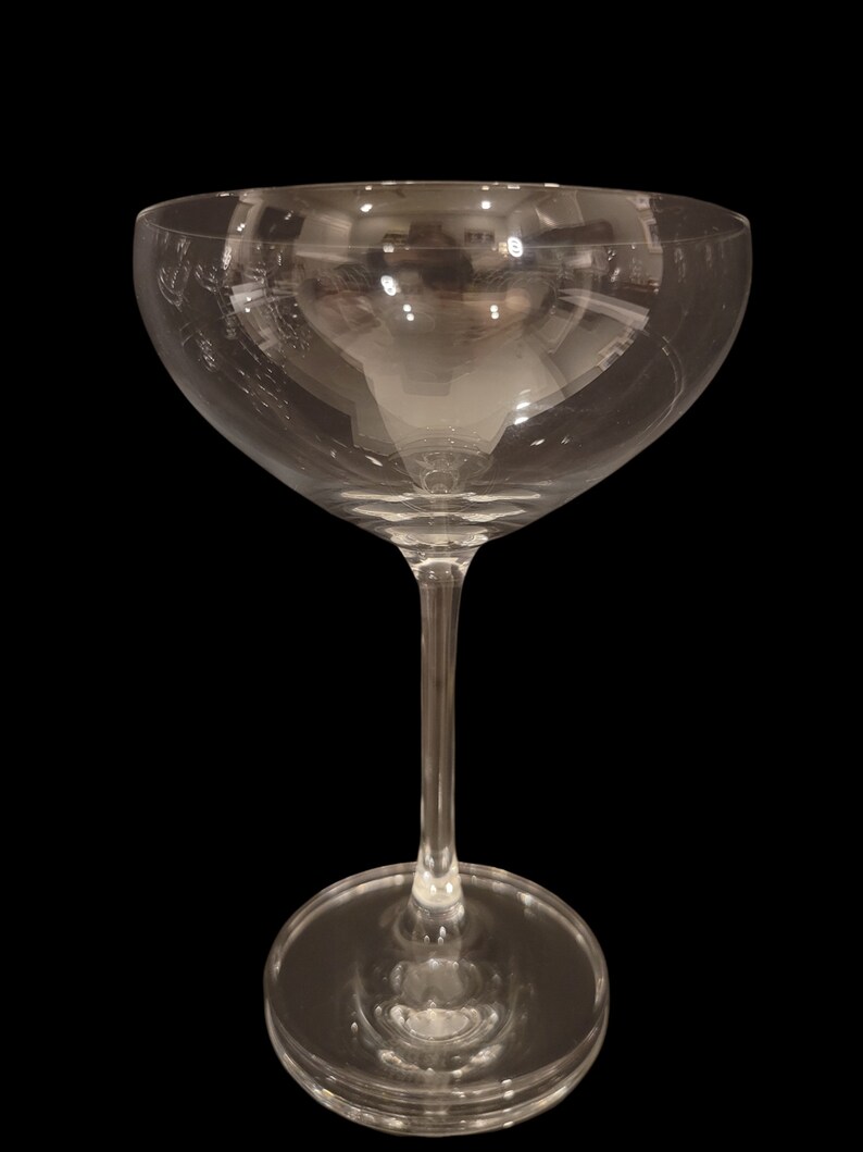 May include: A clear, stemmed champagne coupe glass with a wide, shallow bowl and a thin stem. The glass sits on a round base. The glass reflects the surrounding environment, with a black background.