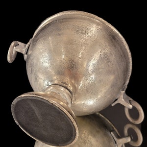 May include: A silver-colored, spherical bowl with two handles and a pedestal base. The bowl has a textured surface and a dark, oval-shaped base. The object is set against a black background, with a reflection visible below.