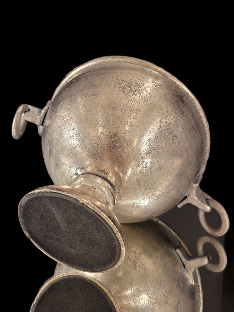 May include: A silver-colored, spherical bowl with two handles and a pedestal base. The bowl has a textured surface and a dark, oval-shaped base. The object is set against a black background, with a reflection visible below.