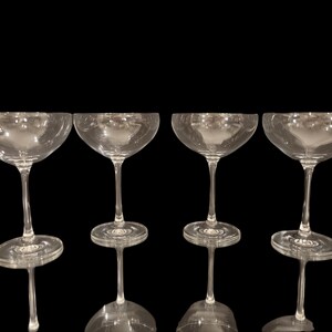 May include: Four clear champagne coupe glasses with a wide, shallow bowl and a long stem. The glasses are arranged in a row against a black background, showcasing their elegant design. The glasses are made of clear glass.