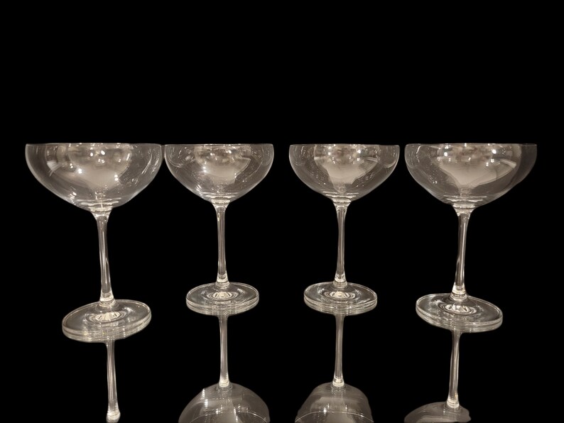 May include: Four clear champagne coupe glasses with a wide, shallow bowl and a long stem. The glasses are arranged in a row against a black background, showcasing their elegant design. The glasses are made of clear glass.