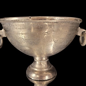 May include: A silver-colored chalice with a textured surface and two ring handles. The chalice has a wide bowl shape and sits atop a pedestal base. The background is black.