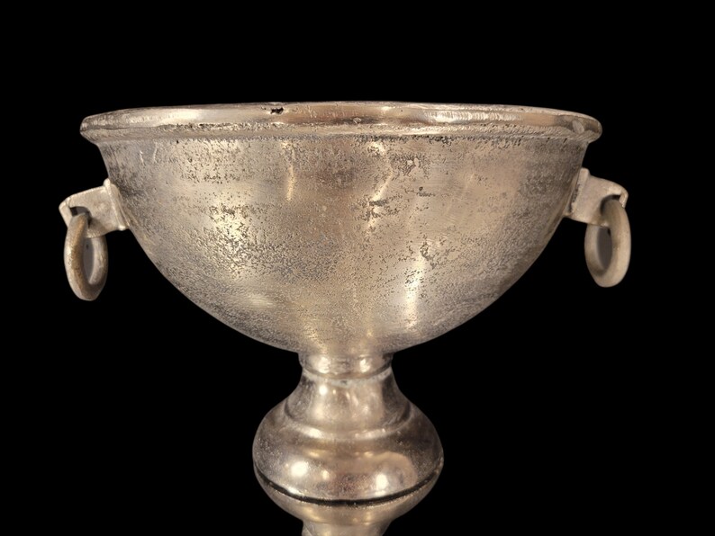 May include: A silver-colored chalice with a textured surface and two ring handles. The chalice has a wide bowl shape and sits atop a pedestal base. The background is black.