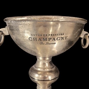 May include: A silver champagne bucket with the words "CUVE&Eacute; DE PRESTIGE CHAMPAGNE Du Louvois" engraved on the front. The bucket has two ring handles and sits on a pedestal base. The background is black.