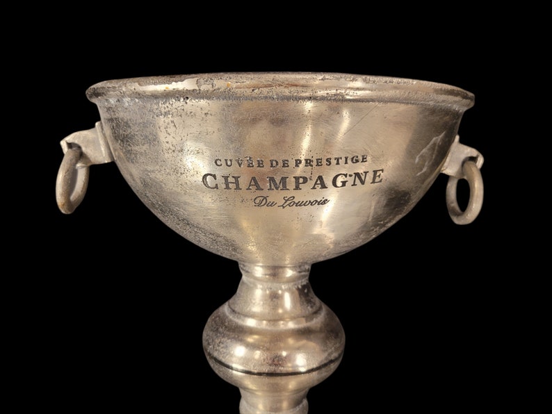 May include: A silver champagne bucket with the words "CUVE&Eacute; DE PRESTIGE CHAMPAGNE Du Louvois" engraved on the front. The bucket has two ring handles and sits on a pedestal base. The background is black.