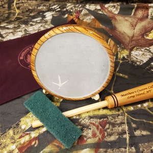May include: A wooden slate call with a gray slate surface and a wooden handle. The slate call is on a wooden stand and has a small green cleaning sponge. The handle has the text "Matthew's Custom Calls Long Island, NY" engraved on it.