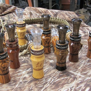 May include: A collection of eight wooden duck calls with a black and brown wood grain finish. Each call has a clear plastic insert and a black band with the text "Mathew's Custom Calls".