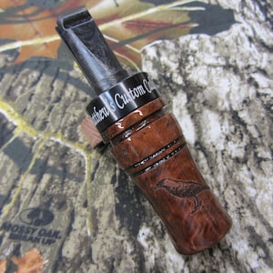 May include: A brown and black wooden duck call with a black metal band engraved with "Matthew's Custom Calls". The call has a carved raven design on the side.