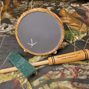 May include: A wooden turkey call with a black and brown striped design. The call has the text "Matthew's Custom Calls Long Island, NY" engraved on the side. The call is accompanied by a slate turkey call with a turkey footprint design and a green cleaning pad.