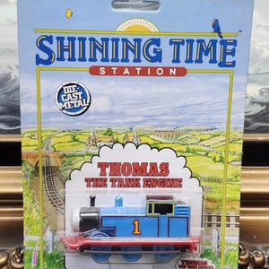 May include: A blue die-cast metal Thomas the Tank Engine toy train in its original packaging. The card features the Shining Time Station logo, a landscape illustration, and the text Thomas the Tank Engine with the number 1 on the engine side.