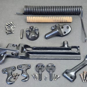 May include: A collection of black metal hardware components including a large tension spring, a wooden dowel, various mounting brackets, a long hinge mechanism, a hook-shaped piece, and several small silver-coloured screws for assembly and installation.