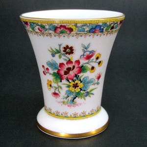 May include: White porcelain vase with a flared shape, decorated with a floral pattern in red, pink, yellow, and blue. It has a gold rim and base. The vase is approximately 15 cm tall.
