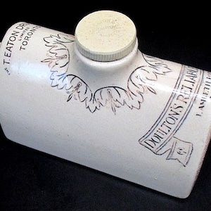 Könnte beinhalten: Cremefarbene, rechteckige Keramik-Wärmflasche mit Schraubverschluss. Mit schwarzem Text und floralen Designs. Der Text lautet "T. Eaton DR. LIMITED TORONTO" und "DOULTON'S RELIABLE LAMBETH LONDON."