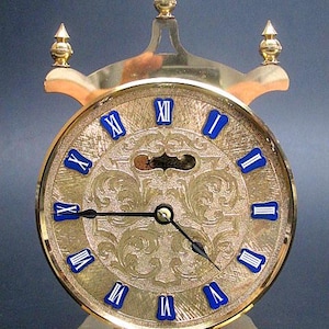Peut inclure: Une horloge dorée ornée avec un cadran rond et un support décoratif. Le cadran de l'horloge présente des chiffres romains bleus et un motif floral détaillé. L'horloge a un cadre doré et deux finials décoratifs.