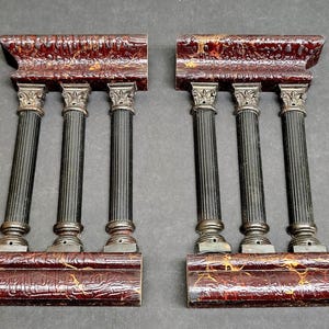 May include: A pair of decorative architectural pieces, each with three black fluted columns and silver-toned capitals. They sit on a textured, reddish-brown base and are topped with a matching cornice. The design has a vintage appearance, reminiscent of classical architecture.