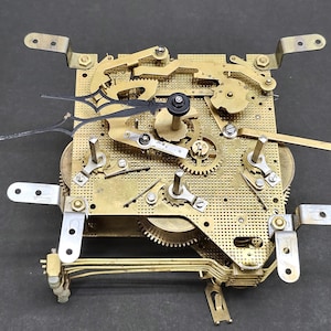 May include: A clock movement featuring brass gears, levers, and mounting brackets. The mechanism has black hands and intricate details, revealing the inner workings of a clock. The design is a complex arrangement of mechanical components, typical of vintage timepieces.