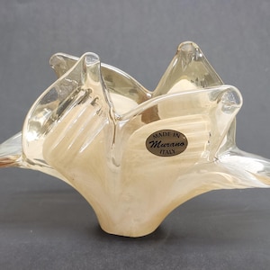 May include: A clear and beige glass bowl with a ruffled, organic shape. The glass features a swirling, translucent pattern and a smooth, glossy finish. A circular sticker on the side displays the text "MADE IN Murano ITALY" in black lettering.