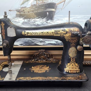 May include: A vintage black Singer sewing machine featuring intricate gold filigree decals and a prominent hand wheel on the right side. The machine body displays the Singer brand name in gold lettering and includes a metallic needle plate.