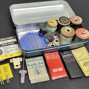 Könnte beinhalten: Eine Nähset-Dose aus Metall mit verschiedenen Holzspulen, Metallspulen und Nadeln. Neben der Dose liegen mehrere alte Nadelpackungen mit den Aufschriften John James, Needle-Pak, Sharps und Milwards sowie ein kleines Kunststoffwerkzeug.