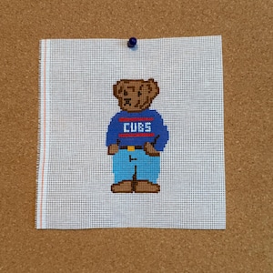 Peut inclure: Une toile de canevas blanche carrée représentant un ours brun brodé au point de croix, vêtu d'un pull bleu avec le mot CUBS en lettres blanches, des rayures rouges et un pantalon bleu clair. Une petite punaise bleue est fixée en haut au centre.