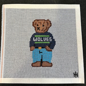 May include: Needlepoint canvas featuring a brown bear wearing a navy blue sweater with the word "WOLVES" in white and green. The bear is also wearing blue jeans. The canvas is white with a grid pattern.