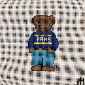 May include: Needlepoint canvas depicting a brown bear in a blue sweater with the word "RAMS" in yellow. The bear wears blue trousers and brown shoes. The background is white with a grid pattern.