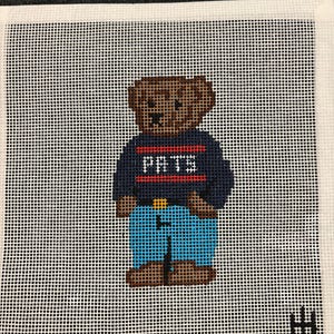 May include: Needlepoint canvas featuring a brown teddy bear wearing a navy blue sweater with the letters "PRTS" in red and white. The bear is also wearing blue jeans with a gold buckle. The background is white with a grid pattern.