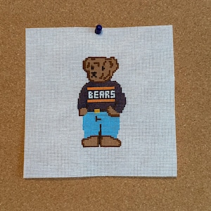 May include: A square piece of white needlepoint canvas featuring a cross-stitched brown bear wearing a dark navy blue sweater with the word BEARS in white letters, orange stripes, and bright blue pants, secured with a single blue push pin.