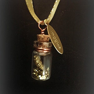 Könnte beinhalten: Eine goldene Halskette mit einer kleinen Glasflasche gefüllt mit goldenem Glitzer und einem goldenen Anhänger mit der Aufschrift "Imagine".