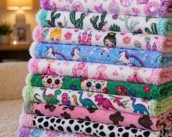 May include: A stack of colorful, plush blankets with various patterns. Designs include unicorns, princesses, dinosaurs, cacti, and cow spots. Each blanket has a contrasting trim, with colors like pink, green, and blue. The blankets are soft and inviting.