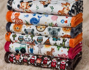 May include: A stack of six plush baby blankets with various animal and themed patterns. The blankets feature designs of foxes, an octopus, a whale, a lion, a koala, owls, cacti, a tractor, and sock monkeys. Each blanket has a different colored trim.