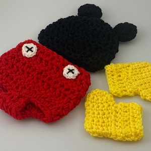 May include: A handmade crochet set featuring a black Mickey Mouse hat with two round ears, red shorts with white and black button accents, and yellow gloves. The set is likely for a baby or toddler.
