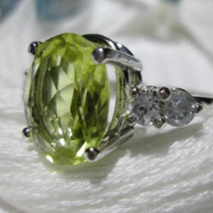 Puede incluir: Un anillo de plata con una piedra preciosa grande de forma ovalada de color verde claro y dos piedras peque&ntilde;as redondas transparentes.