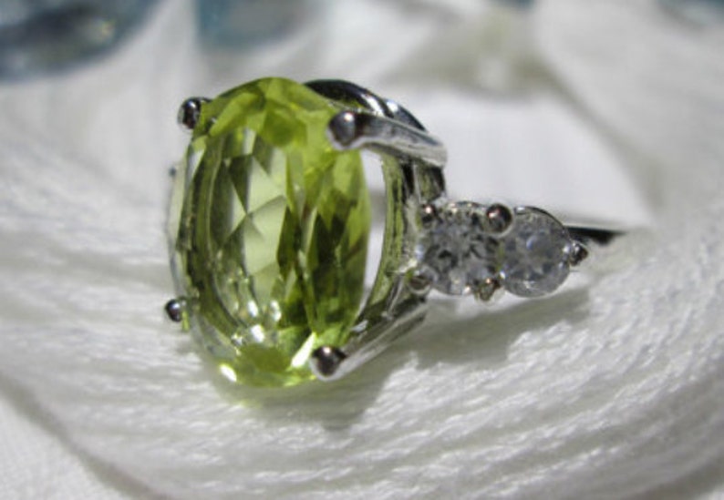 Puede incluir: Un anillo de plata con una piedra preciosa grande de forma ovalada de color verde claro y dos piedras peque&ntilde;as redondas transparentes.