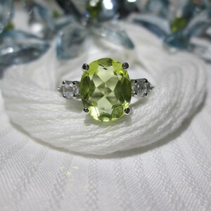 Puede incluir: Un anillo de plata con una piedra preciosa verde grande de forma ovalada y dos piedras preciosas redondas y transparentes m&aacute;s peque&ntilde;as en la banda.