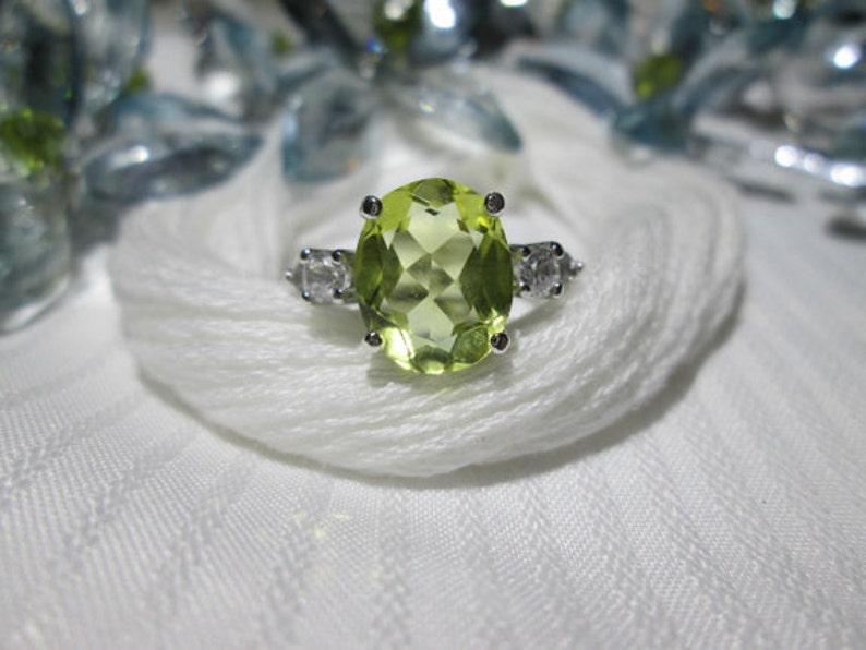 Puede incluir: Un anillo de plata con una piedra preciosa verde grande de forma ovalada y dos piedras preciosas redondas y transparentes m&aacute;s peque&ntilde;as en la banda.