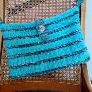 Puede incluir: Un bolso tejido azul turquesa con un patrón a rayas y un cierre de botón plateado. El bolso está sentado en una silla de madera con un asiento de caña tejido.