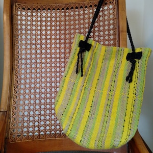 May include: A yellow and green striped woven bag with black tassels hanging from the top. The bag is sitting on a wooden chair with a woven cane seat.