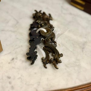 May include: A collection of dark bronze-toned metal drawer pulls featuring intricate scrollwork and ornate detailing. The hardware pieces are stacked together and secured with a thin white ribbon threaded through the mounting holes.