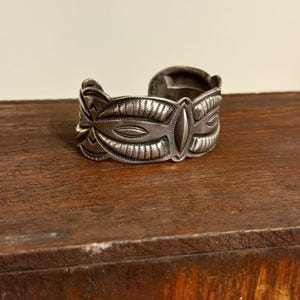 May include: A silver cuff bracelet featuring an oxidized finish and intricate hand-stamped geometric patterns. The metal surface displays a series of repeating oval and chevron motifs with fine line detailing across the entire outer band.