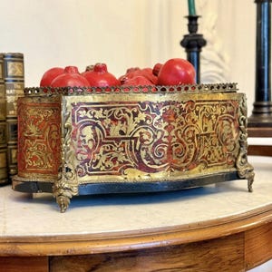 May include: A rectangular decorative box featuring ornate gold-toned metal trim and feet with a red and gold floral pattern on the sides. The top edge has a scalloped metal border, and the interior is filled with several round red pomegranates.