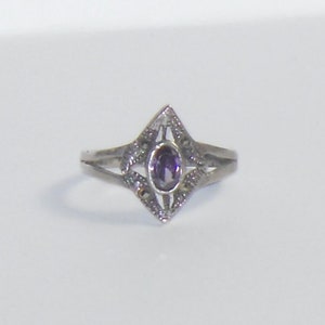 May include: A silver ring with an oval, purple gemstone set in a diamond-shaped frame. The ring has a delicate, openwork design with small, decorative accents around the frame and on the band.