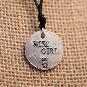 Puede incluir: Un collar de plata con un colgante redondo que dice "WISE GIRL" y tiene un pequeño diseño de búho. El collar está en un cordón negro.