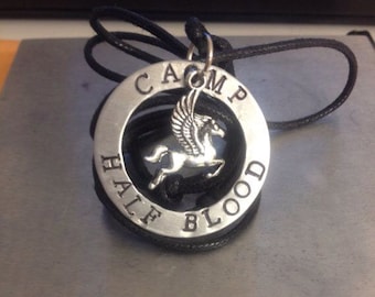 Pode incluir: Um colar de prata com um pingente redondo que diz "Camp Half-Blood" com um cavalo alado dentro do círculo.