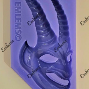 May include: Purple silicone mold with a detailed design of a horned creature's face. The mold is made by Emlems.