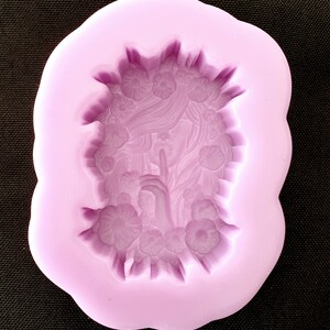 May include: Purple silicone mold with a detailed design of a woman's face with flowers surrounding her. The woman is holding her finger to her lips as if to say "shhh".